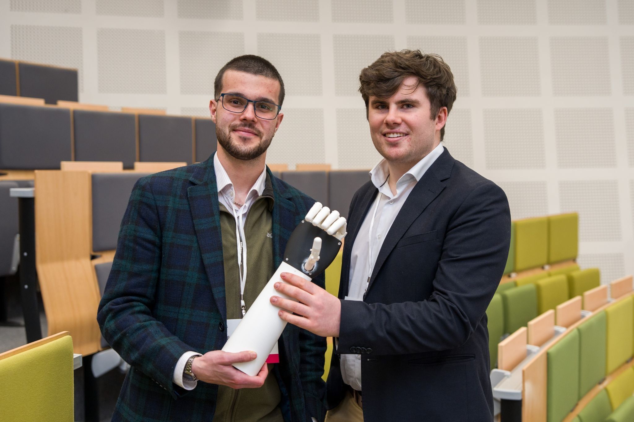 Seán Barrett and William Hartley in a lecture theatre at UCD, holding a white robotic hand between them.